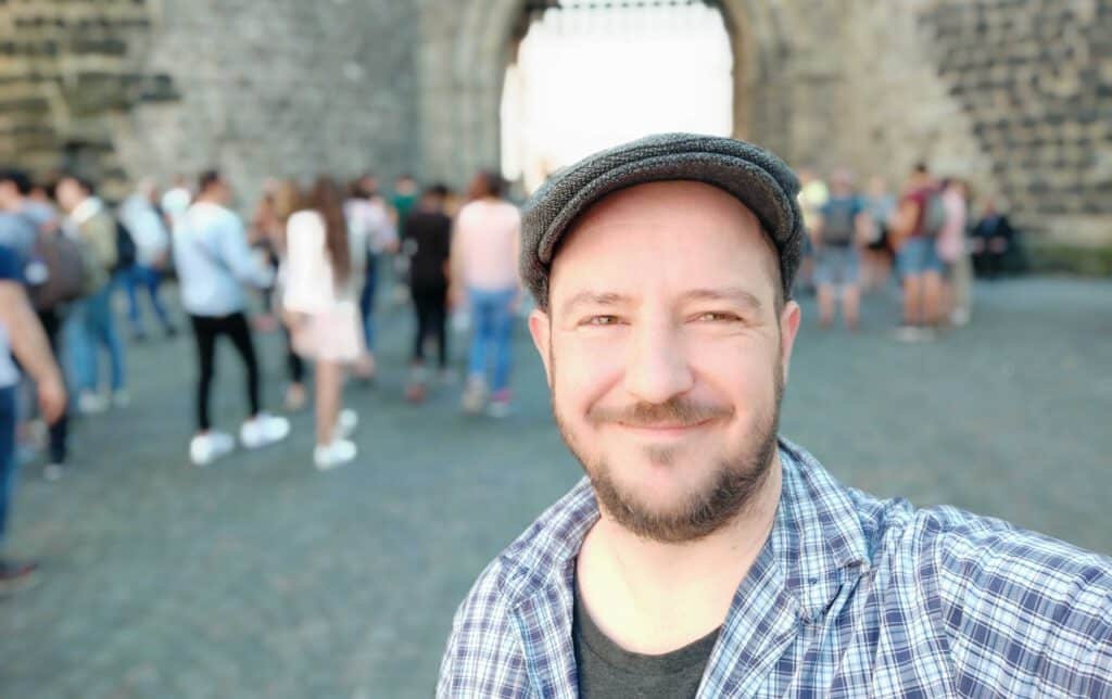 Un hombre con camisa de cuadros y gorra plana sonríe para hacerse un selfie delante de una antigua puerta de piedra durante un recorrido por Colonia a pie, con un grupo de personas difuminadas al fondo.
