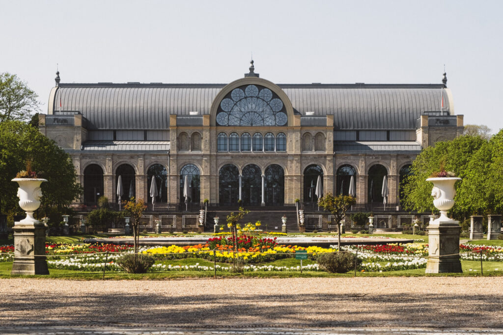 Gran edificio histórico de cristal y piedra con ventanas arqueadas, fachada ornamentada y tejado abovedado, situado detrás de coloridos jardines de flores -una parada emblemática en un recorrido por Colonia a pie- flanqueado por grandes urnas blancas y árboles verdes.