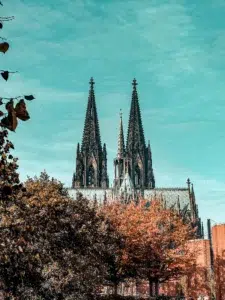 Gothic cathedral with two tall, pointed spires rises above autumn-colored trees under a blue sky, a stunning sight often admired during a freewalk cologne tour.