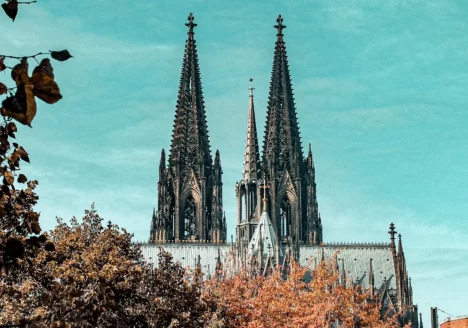 Gothic cathedral with two tall, pointed spires rises above autumn-colored trees under a blue sky, a stunning sight often admired during a freewalk cologne tour.