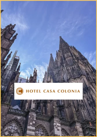 17 Gothic cathedral spires reach into a blue sky, with scaffolding on the left. Overlay text displays the logo and name Hotel Casa Colonia, inviting guests to explore with a freewalk Cologne adventure nearby.